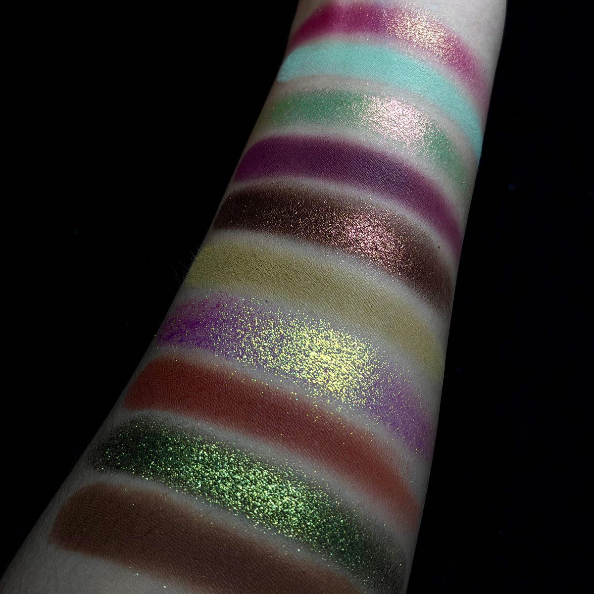 A close-up of a forearm with horizontal swatches of shimmering eyeshadow or pigment in multiple colors, including pink, aqua, green, mauve, bronze, gold, purple, red, olive green, and brown, against a black background.