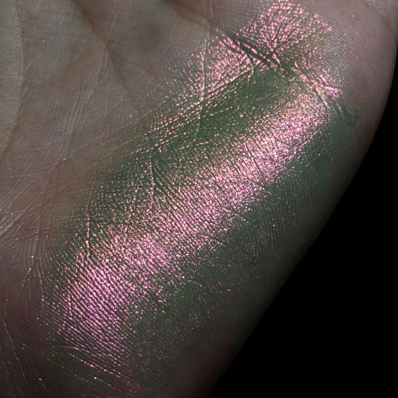 A close-up of a hand showing a shimmering metallic swatch of product applied across the palm, with a pink-lilac cast and green undertone against a black background.