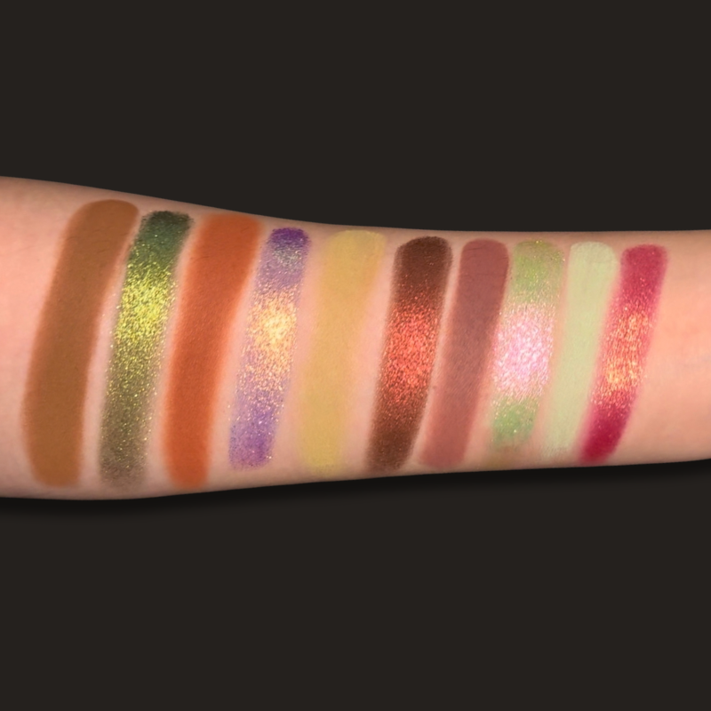 A close-up of a forearm swatched with eight vertical makeup color bars in shimmering shades including brown, olive green, terracotta orange, lavender purple, pale yellow, copper red, mauve pink, mint green, and bright pink, set against a solid black background.