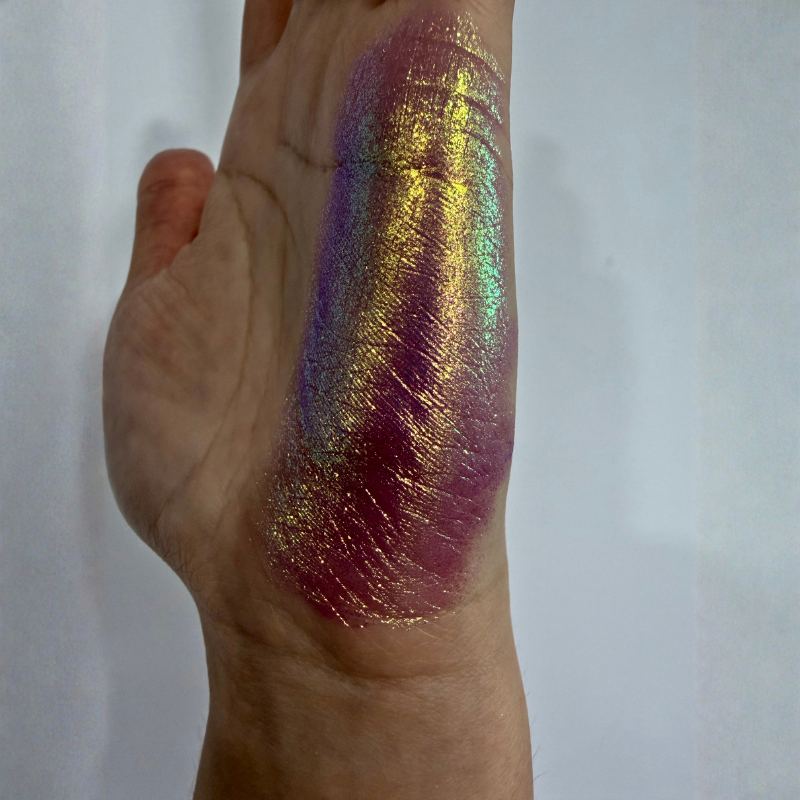 A close-up of a hand showing a thick, shimmery cosmetic swatch on the back of the hand. The product has an iridescent, holographic finish shifting between gold, purple, pink, and green, applied in a thick vertical streak.