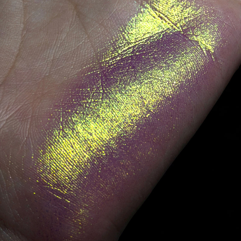 A close-up of a hand showing a shimmering metallic swatch of product, with a bright yellow-green reflective finish over a purple base, against a black background.