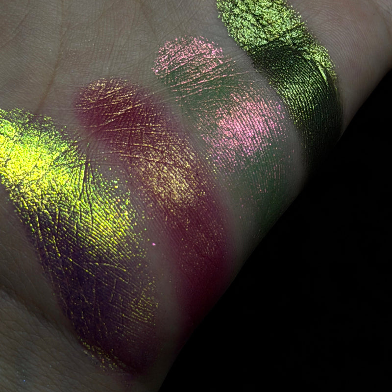 Four metallic eyeshadow swatches in neon yellow, magenta, pink, and olive green applied on the back of a hand against a black background.
